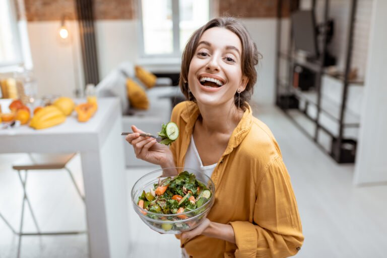 Portrait of a young and cheerful woman dressed in bright shirt eating salad at home. Concept of wellbeing, healthy food and homeliness